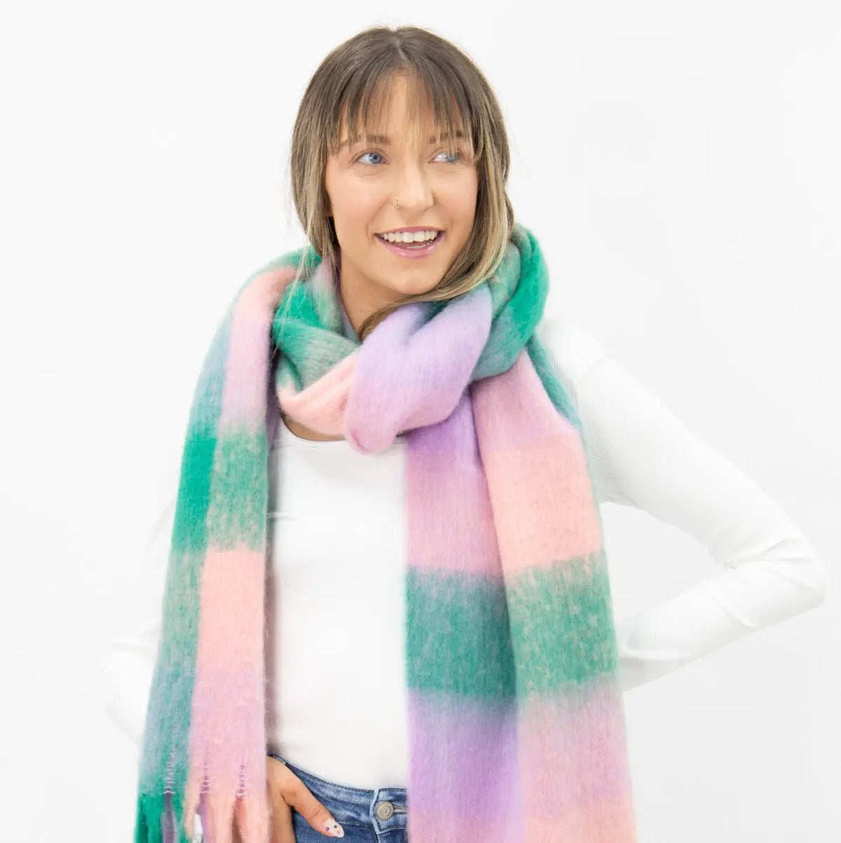Woman wearing an oversized brushed woven plaid scarf in pastel pink, lavender purple and green with long fringe, styled with a white sweater and jeans
