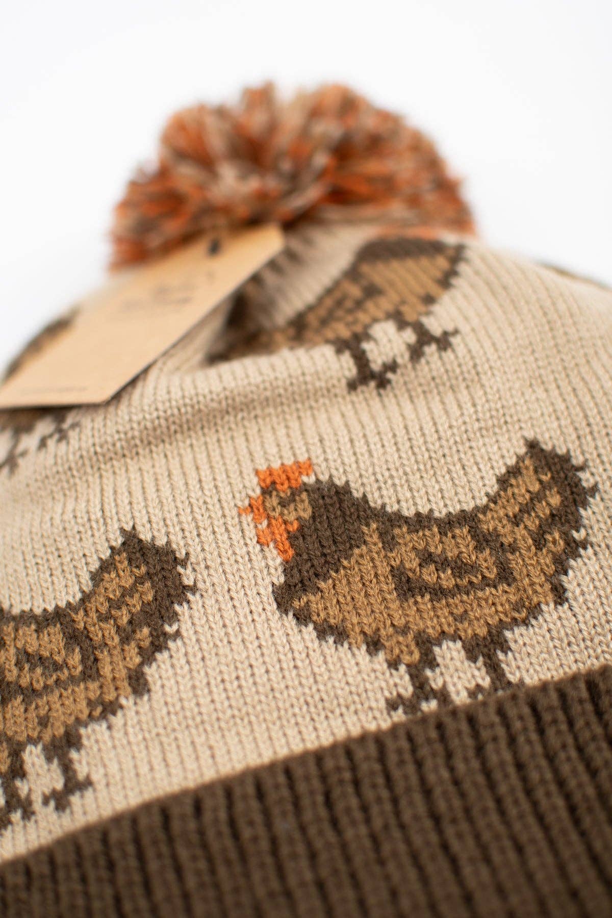 Close-up of brown hen knit pattern with rust-orange accents on an oatmeal farmhouse winter beanie made from 100% acrylic.