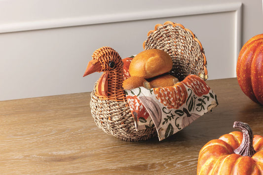 Seagrass turkey-shaped Thanksgiving basket filled with dinner rolls and decorated with an autumn floral kitchen towel on a fall table.