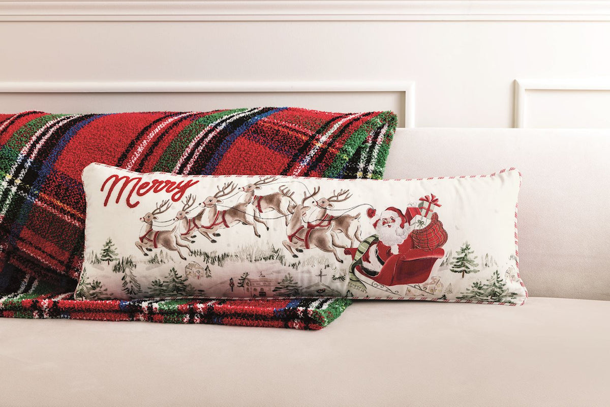 Mud Pie Santa Scene 35-inch Christmas lumbar pillow featuring Santa's sleigh and reindeer, styled on a cream sofa with a red and green plaid blanket.