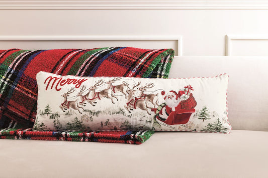 Mud Pie Santa Scene 35-inch Christmas lumbar pillow featuring Santa's sleigh and reindeer, styled on a cream sofa with a red and green plaid blanket.